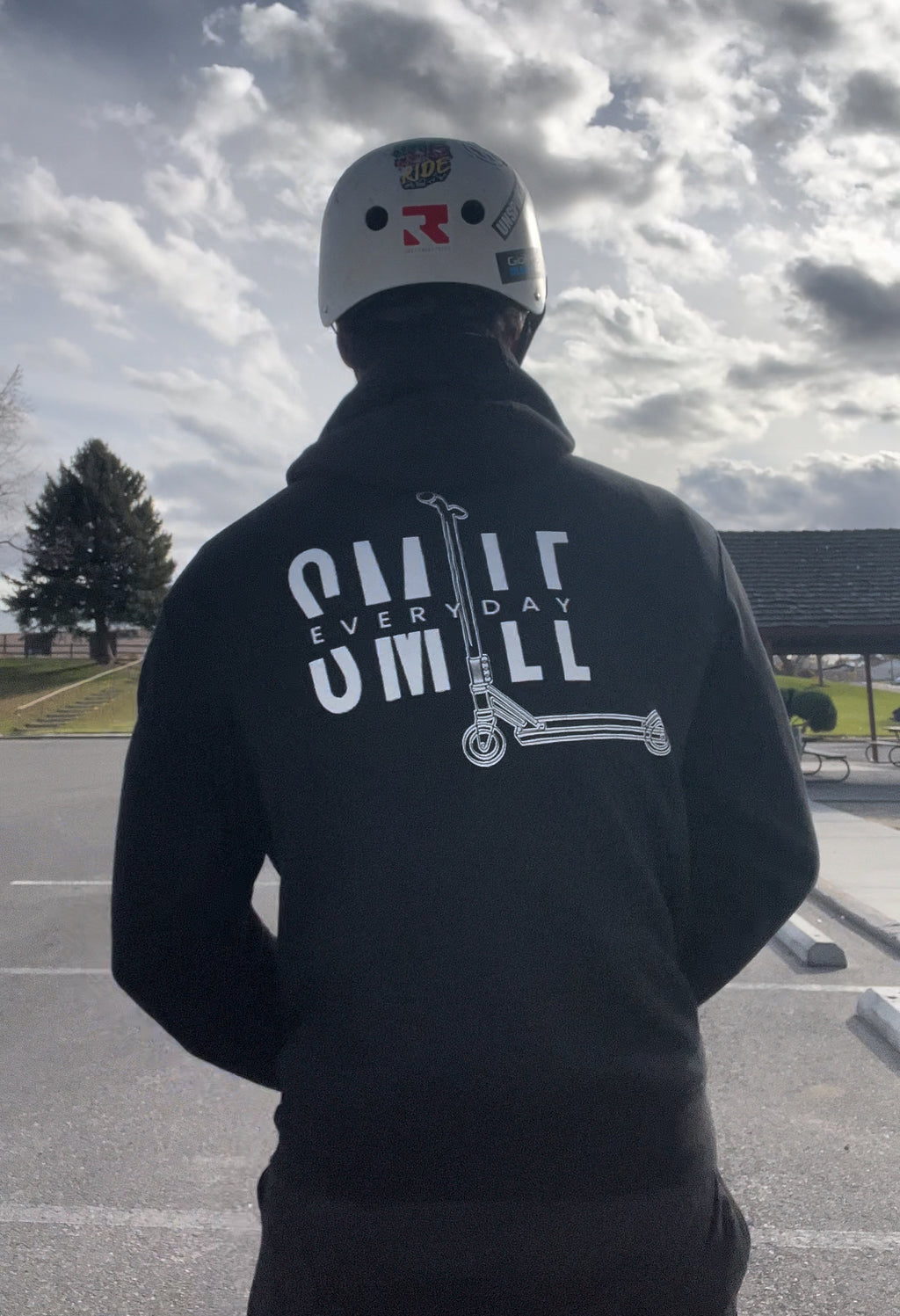 Person wearing a black hoodie with text and graphics, standing outdoors on a cloudy day.
