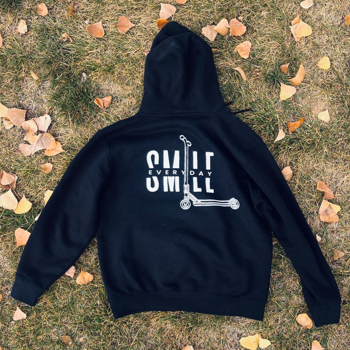 Black hoodie with 'Smile Smile' design on grass with fallen leaves